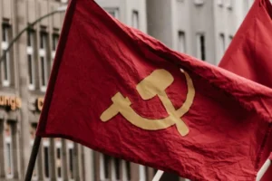 Bandeira comunista vermelha tremulando em destaque, exibindo o símbolo clássico da foice e martelo em amarelo no centro, com um fundo urbano desfocado, representando como começar a estudar comunismo.