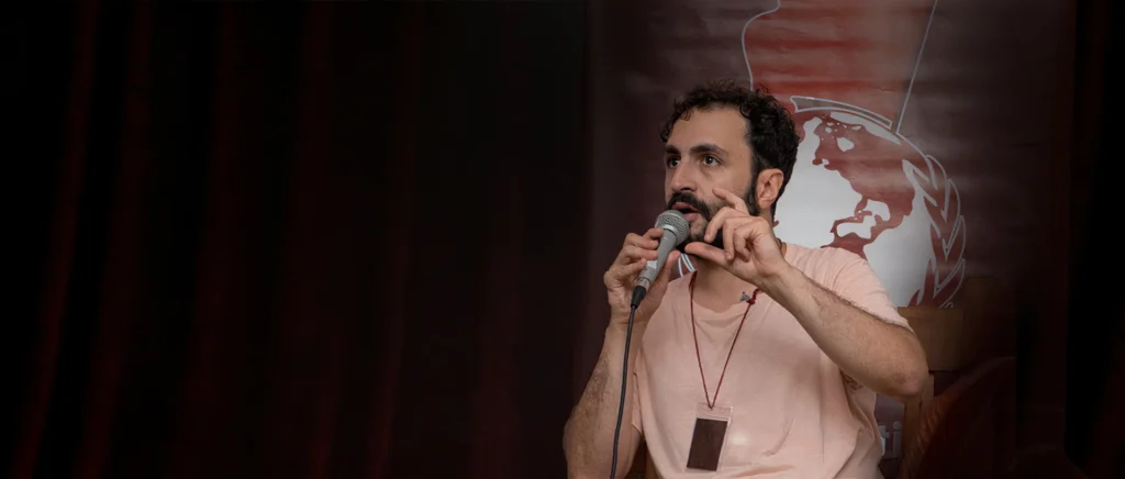 Retrato do professor e historiador Gustavo Gaiofato com um microfone na mão, palestrando, e ao fundo uma cortina vermelha, com um globo terrestre à direita, atrás do professor.