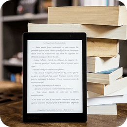 E-reader ligado, encostado numa pilha de livros sobre uma mesa e ao fundo uma estante de livros.