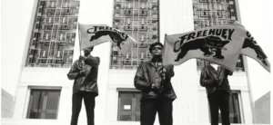 Três militantes dos Panteras Negras uniformizados nas escadarias de um tribunal segurando bandeiras com o logo da pantera e a frase 'Free Huey'.