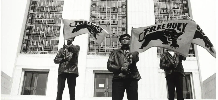 Três militantes dos Panteras Negras uniformizados nas escadarias de um tribunal segurando bandeiras com o logo da pantera e a frase 'Free Huey'.