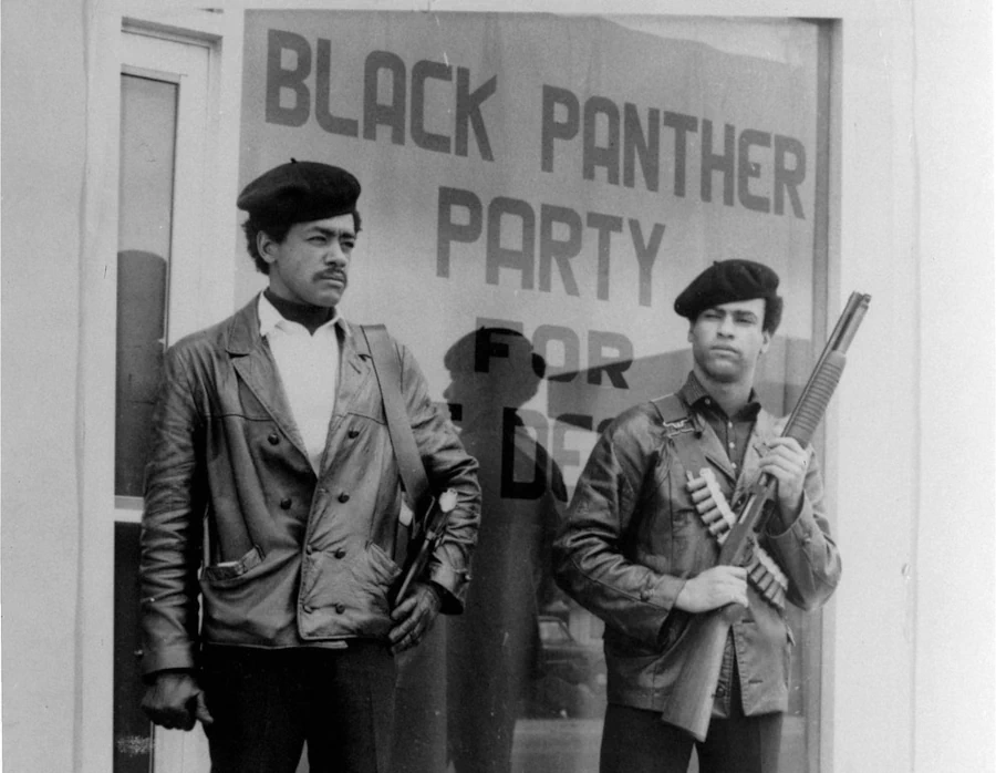Bobby Seale à esquerda e Huey Newton à direita, fundadores do Partido dos Panteras Negras, armados com espingardas, fazendo a segurança em frente à sede da organização sob a placa 'Black Panther Party for Self Defense'.