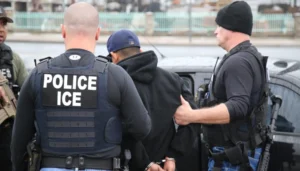Agente da polícia com colete escrito Police ICE nos EUA detém um homem algemado ao lado de um veículo durante uma operação de fiscalização.