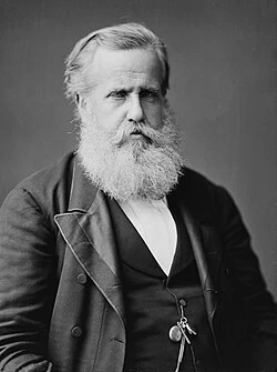 Uma fotografia histórica em preto e branco (retrato de estúdio) mostrando o Imperador Dom Pedro II do Brasil na velhice. Ele tem uma barba branca longa e cheia e cabelos brancos, olhando diretamente para a câmera com uma expressão séria. Ele veste um paletó escuro de abotoamento duplo sobre um colete escuro e uma camisa branca com colarinho alto. Uma corrente de relógio de bolso com fobs é visível pendurada em seu colete. O fundo é um estúdio neutro e suave. Esta imagem é representativa do líder do Brasil Império.
