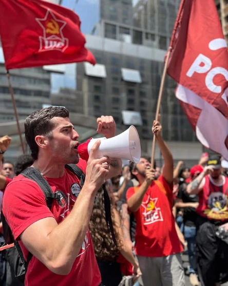 Professor do curso de Introdução à Sociologia, Gabriel Tavares, com um megafone na mão em uma manifestação popular. Atrás dele, há pessoas e duas bandeiras: uma da UJC e outra do PCBR.