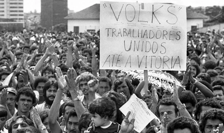 Fotografia em preto e branco de uma grande manifestação de trabalhadores no ABC Paulista. Diversos operários com as mãos levantadas, segurando um cartaz que diz: "'VOLKS' TRABALHADORES UNIDOS ATÉ A VITÓRIA". A imagem captura a mobilização da classe trabalhadora durante as greves de 1978-1980, onde a redução da jornada de trabalho era uma pauta central ao lado de melhores salários e condições.