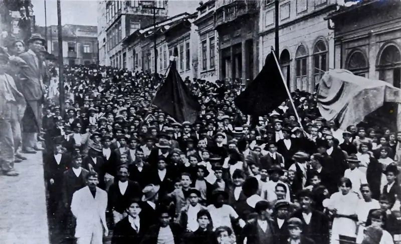 Fotografia histórica em preto e branco da Greve Geral de 1917 em São Paulo. Uma enorme multidão de trabalhadores preenche uma rua íngreme e estreita, ladeada por casarões antigos. No centro, destacam-se duas grandes bandeiras pretas erguidas, simbolizando a organização anarquista e socialista na luta histórica pela redução da jornada de trabalho no Brasil.
