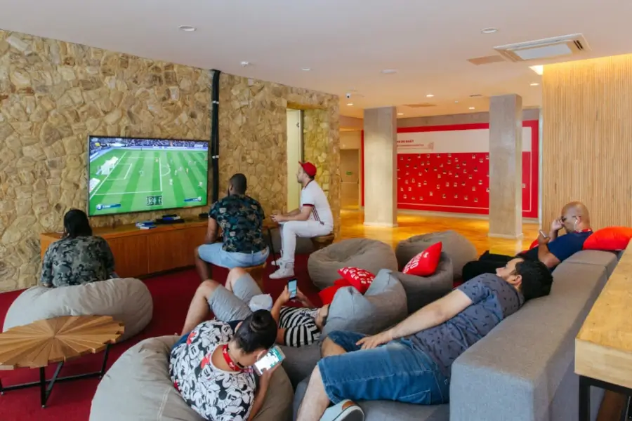 Sala de jogos da sede do iFood em Osasco, SP, com sofás, puffs e um videogame ligado em um jogo de futebol. Um dos funcionários está jogando, enquanto os outros seis funcionários estão relaxando nos sofás e nos puffs. A imagem escancara que a uberização serve para explorar os motoristas e entregadores enquanto a plataforma gera lucros exponenciais.