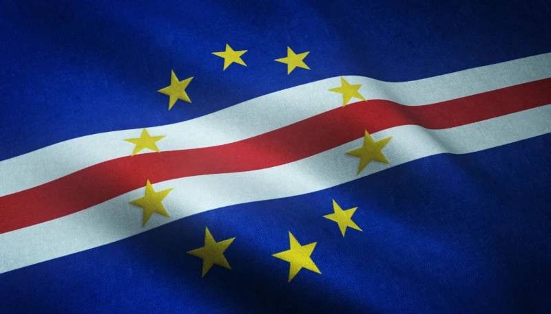 Bandeira oficial de Cabo Verde ondulando em tecido, com fundo azul escuro, três faixas horizontais centrais nas cores branca, vermelha e branca, e um círculo com dez estrelas amarelas de cinco pontas.