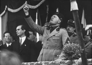 Benito Mussolini fazendo um discurso eloquente, com outros homens e generais ao redor, representando o que é o fascismo. Fonte da imagem: https://www.worldhistory.org/trans/pt/1-19718/benito-mussolini/